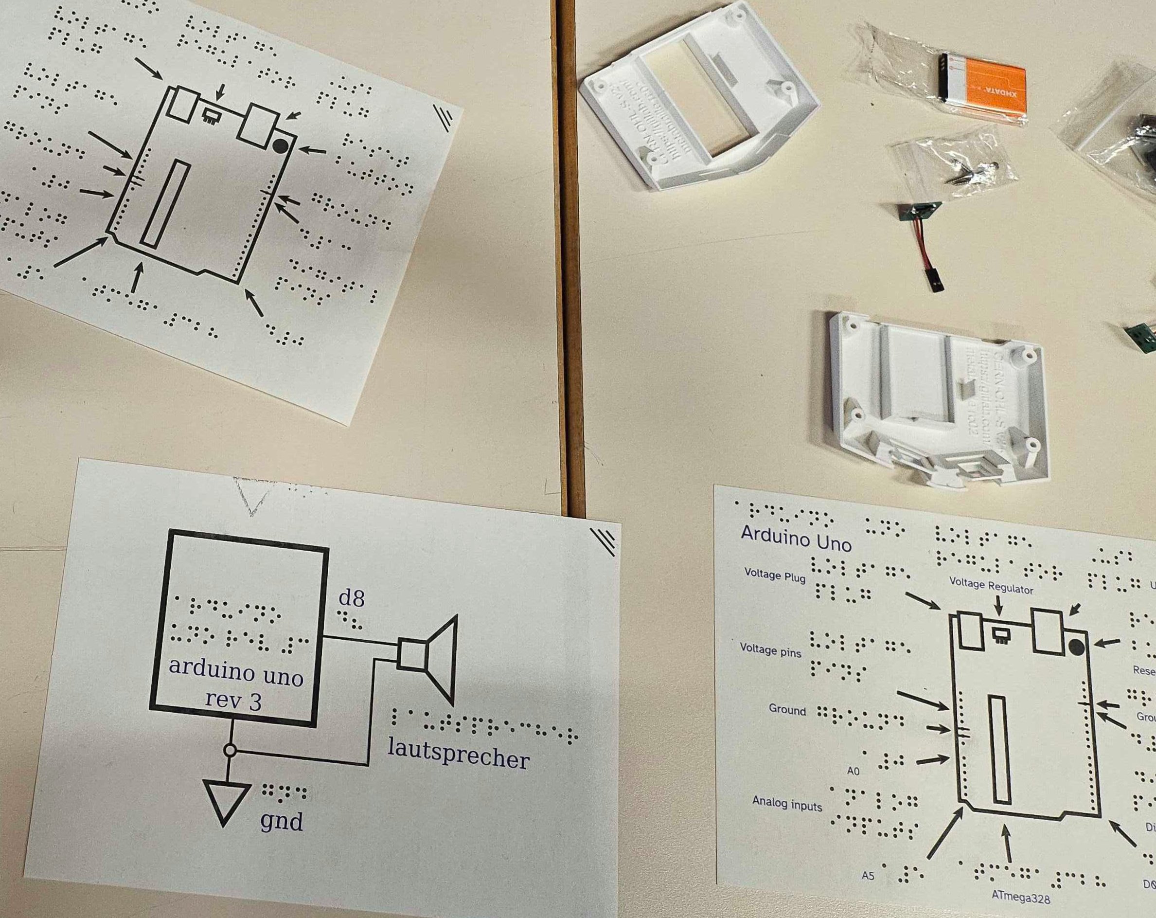 3 plans for the keyboard construction lie on the table. Each one is labeled with English and Braille. In the top right corner is a keyboard shell, battery pack and key-parts.